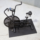 A black Endurance Pro indoor exercise bike with a digital display screen, mounted on a black rectangular mat on a concrete floor.