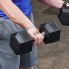 A close-up lifestyle shot of a person lifting a black hex dumbbell in a gym setting.
