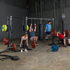 A group of people working out in a gym with a large black steel power rack. One person is hanging from the pull-up bar, another is lifting a barbell, a third is standing nearby, and another is doing a bench press.