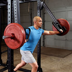 A man in a blue t-shirt and gray shorts uses a bodyweight pull-up bar on a power rack with red weight plates in a garage setting.