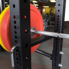 A close-up of a black adjustable weight bench or squat rack with a loaded barbell in a gym setting. Red and yellow weight plates are visible on the left, and the bench frame has measurement markings.